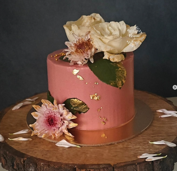 Pink floral celebration cake with gold leaf on rustic wood slice - Vivante Patisserie Bangalore